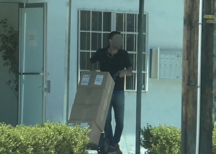 Surveillance image showing a person transporting boxed products with a hand truck outside a business.